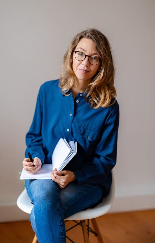 beziehungscoaching-portrait-mirja-schaeffler-hentschel Mirja Schäffler-Hentschel Beziehungscoaching – sitzend auf einem weißen Stuhl mit Holzbeinen, ein Notizbuch und Stift in der Hand.