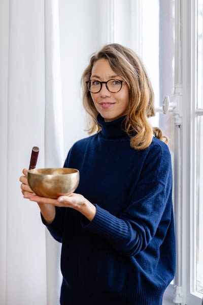 Mirja Schäffler-Hentschel Coach für Persönlichkeitsentwicklung  – stehend am weißen Fenster eine goldene Klagschale in der linken Hand, in der rechten Hand einen dunkelbraunen Klöppel.