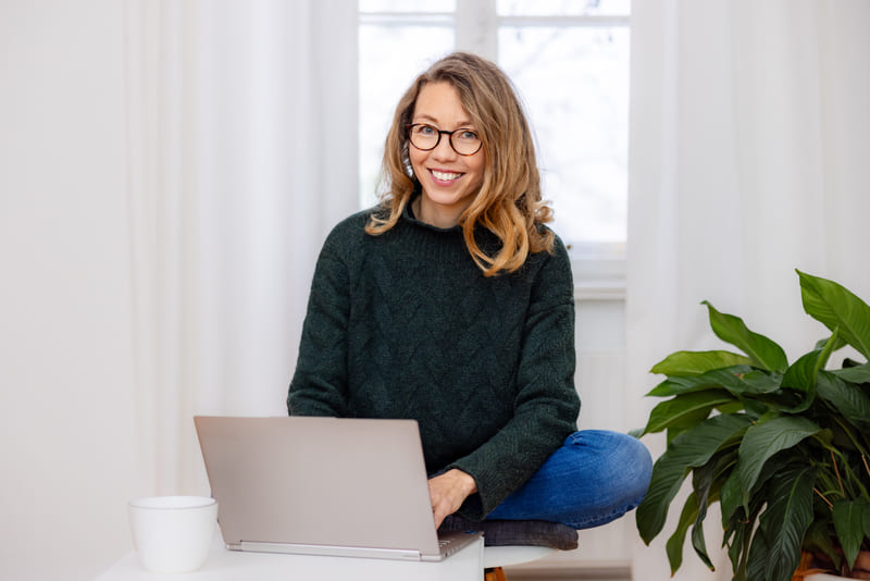 Mirja Schäffler-Hentschel Coach für Persönlichkeitsentwicklung  – sitzend im Schneidersitz auf einem Stuhl, vor einem silbernen Laptop, der auf einem kleinen weißen Tisch steht mit einer weißen Tasse darauf, im Hintergrund ein helles weißes Fenster mit weißen Gardinen, rechts eine grüne Pflanze.
