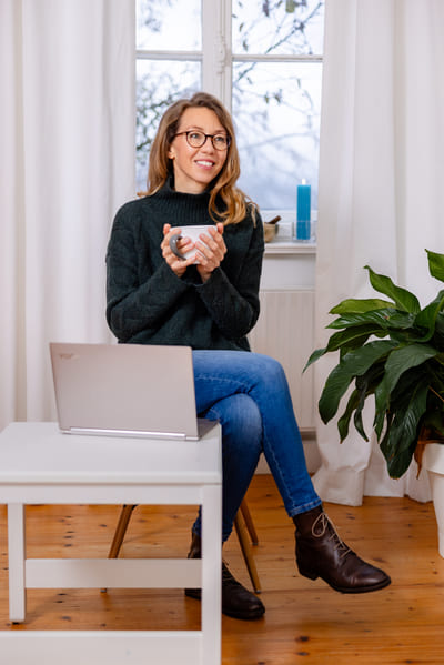 Mirja Schäffler-Hentschel Coach für Persönlichkeitsentwicklung  – sitzend auf einem Stuhl mit Tasse in der Hand, davor ein kleiner weißer Tisch mit silbernem Laptop, im Hintergrund ein weißes helles Fenster mit weißen Gardinen.