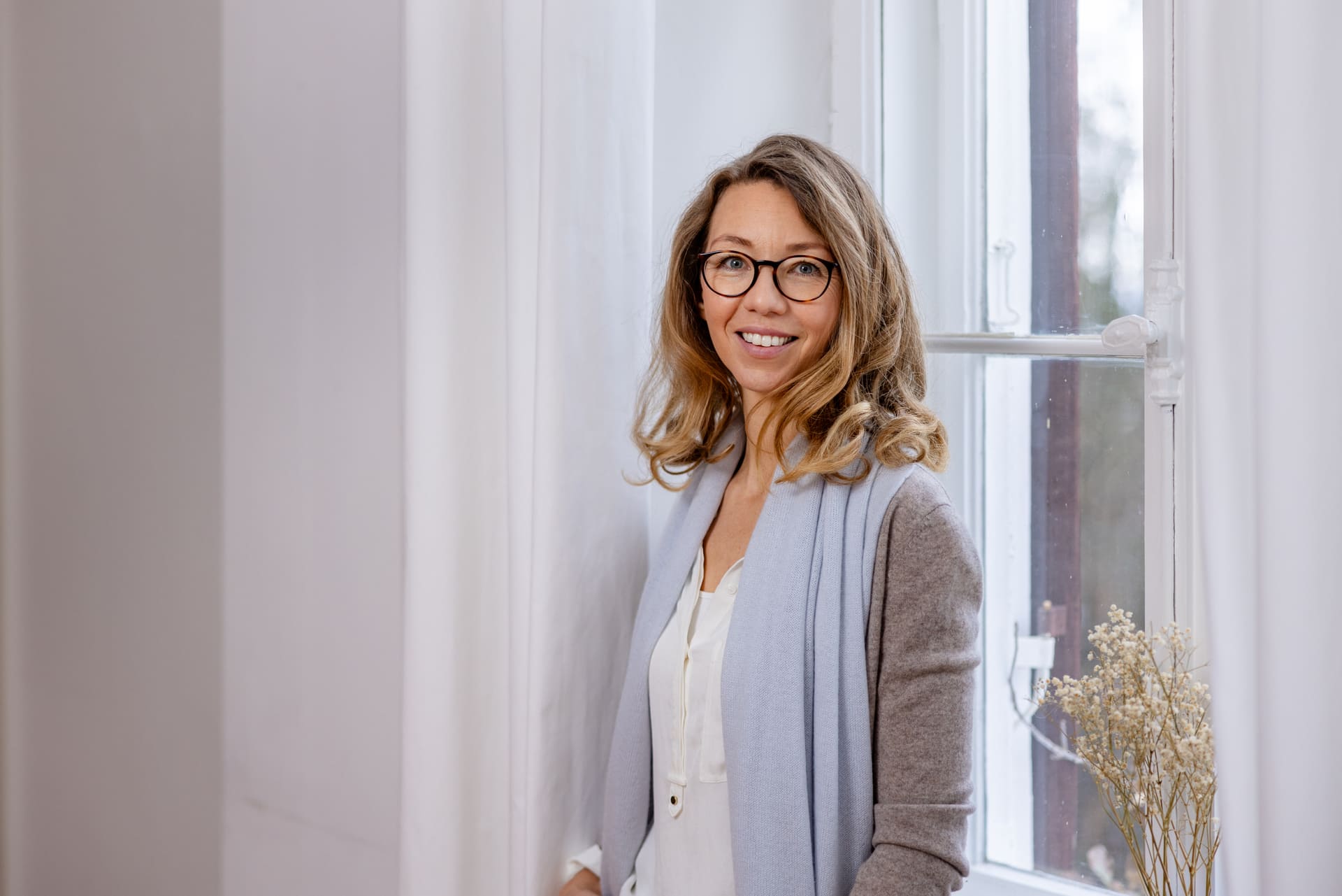 Mirja Schäffler-Hentschel Coach für Persönlichkeitsentwicklung – stehend und leicht angelehnt am hellen weißen Fenster mit weißen Gardinen.