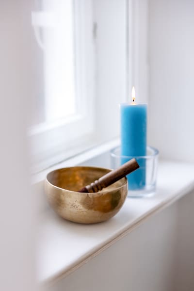 Mirja Schäffler-Hentschel Coaching-Methoden – auf einer weißen Fensterbank steht eine angezündete Kerze in einem kräftigen Blauton in einem Glasbehälter, daneben steht eine goldene Klangschale, aus der ein dunkelbrauner Klöppel herausragt.