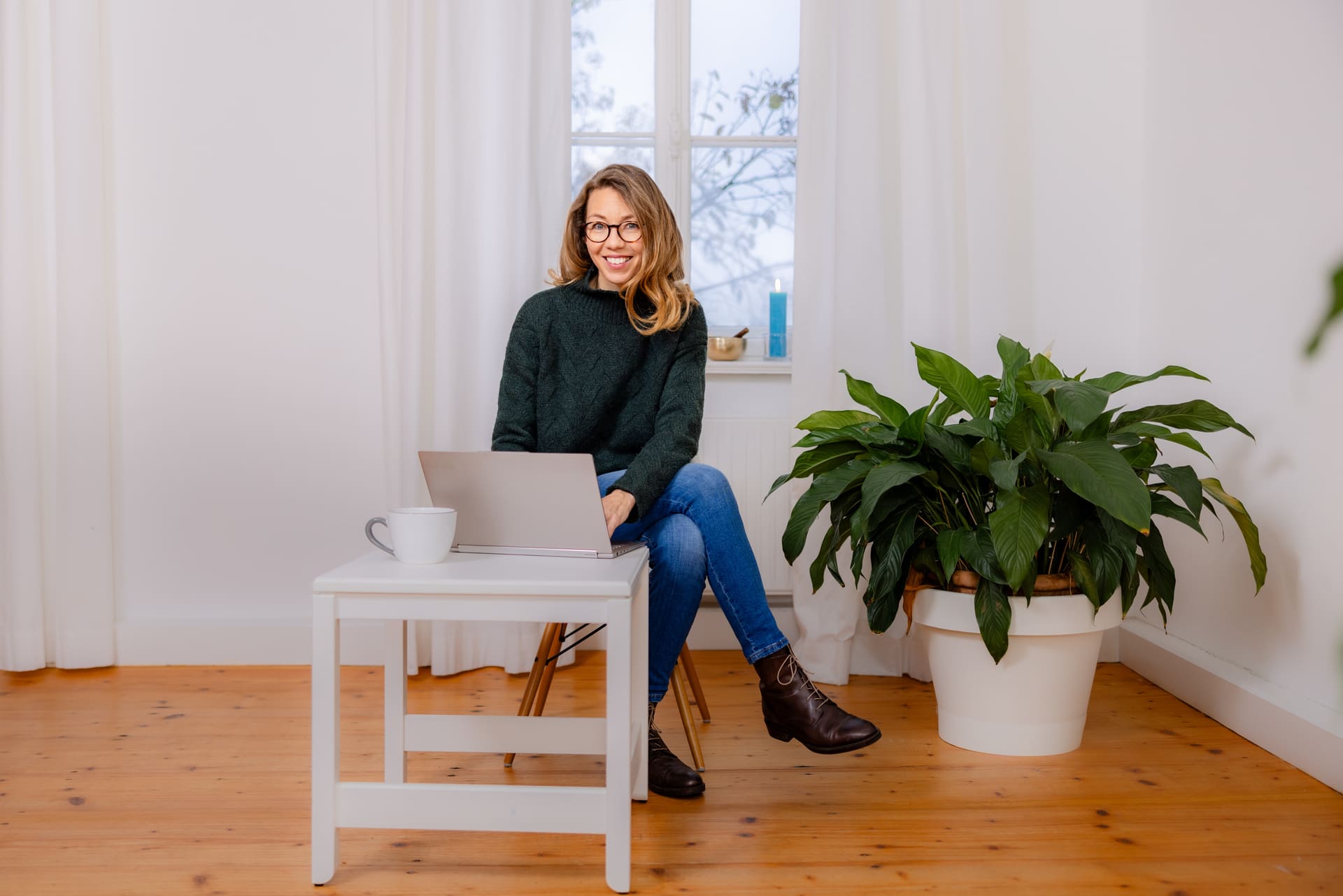 Mirja Schäffler-Hentschel Coaching und Beratung – sitzend, die Beine übereinandergeschlagen auf einem Stuhl, vor einem silbernen Laptop, der auf einem kleinen weißen Tisch steht mit einer weißen Tasse darauf, im Hintergrund ein helles weißes Fenster mit weißen Gardinen und eine grüne Pflanze im weißen Blumentopf.